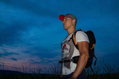 Man with backpack looking away while standing on field against sky during sunset