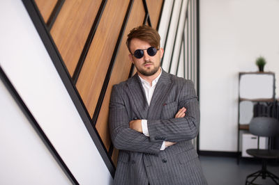 A young man in a suit and sunglasses is standing near the wall with his hands folded