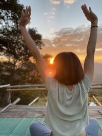 Rear view of woman with arms raised against sky during sunset