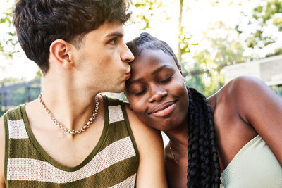 Stylish young male looking away and kissing forehead of happy african american girlfriend during romantic date on sunny summer day on street