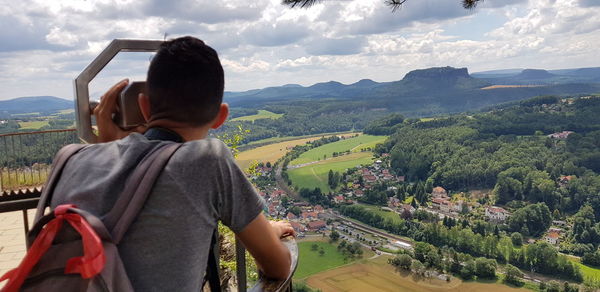 Rear view of young man looking landscape through coin-operated binoculars against cloudy sky