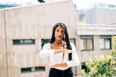 Portrait of young woman gesturing equal sign while standing against building