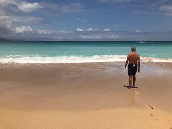 Rear view of shirtless man walking at beach against cloudy sky during sunny day