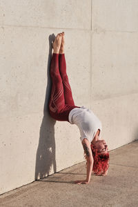 Side view of slim barefooted female with red hair in sportswear standing upside down in downward facing dog pose leaning on concrete wall while training alone on street