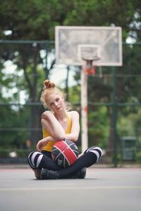 Portrait of young woman sitting with basketball at sports court 