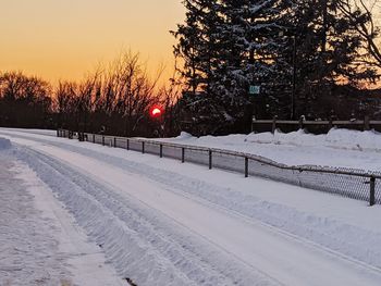 Snow covered road by trees against sky during sunset