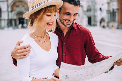 Cheerful young couple reading map while standing in city