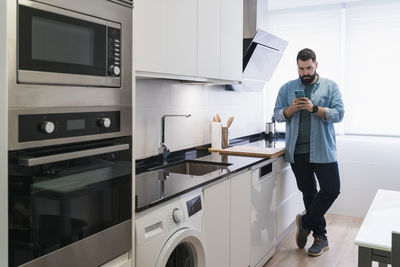 Man cooking crepes in the kitchen with a mobile phone in a denim shirt
