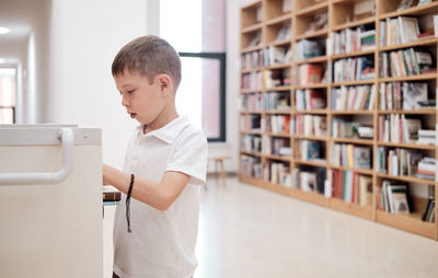 A schoolboy in a white t-shirt puts books in a cart in the library, copy space. 