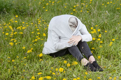 Young woman covering head while sitting on grass by yellow flowering plants