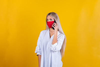 Portrait of woman wearing mask talking over mobile phone against yellow background