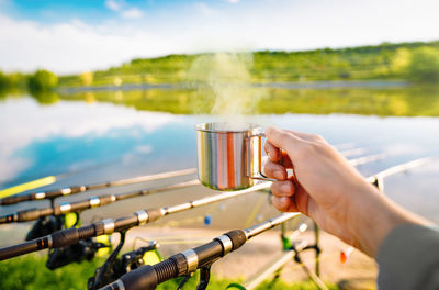 Person drinking hot tea by fishing reels on lakeshore