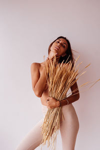 Young shirtless woman holding plants standing by wall