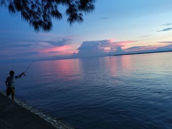 Man fishing in sea against sky during sunset