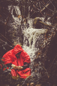 Man wearing red raincoat while sitting against waterfall forest