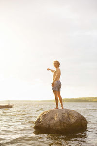 Full length side view of man pointing while standing on rock in lake against sky