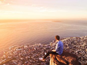 Side view of man sitting on rock over cityscape and seascape during sunset