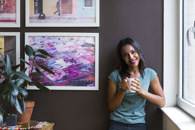 Portrait of woman holding coffee cup while standing against picture frames on wall at home
