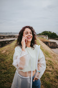 Smiling woman talking over phone while standing on grass against sky