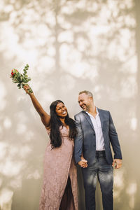 Happy bride holding bouquet with hand raised standing by groom against wall