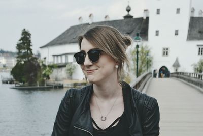 Young woman wearing sunglasses standing against river and footbridge in city