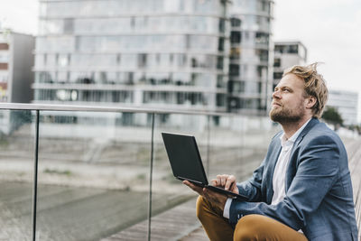 Businessman sitting on bridge using laptop looking up