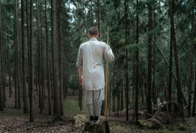 Back view of unrecognizable man with stick standing on rock near trees while practicing kung fu in coniferous forest