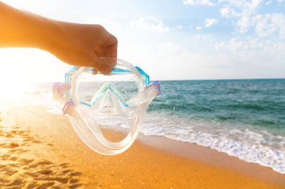 Hand holds snorkeling goggles against the background of the beach and sky. a man's hand holds a pipe