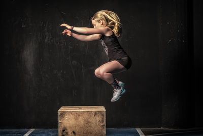 Side view of strong lady in sportswear jumping on shabby wooden box during intense workout in gym