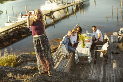 Woman taking selfie through smart phone with friends sitting in background at harbor