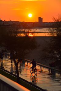 Silhouette man riding bicycle on bridge over river against sky during sunset