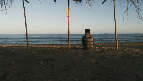 Rear view of man sitting at beach against sky during sunset