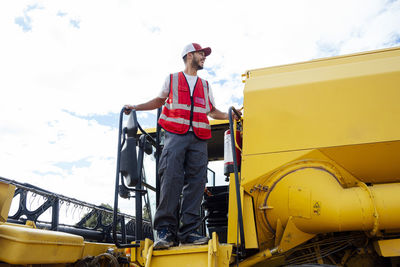 From below full body side of pensive male combine operator in uniform standing on ladder of professional machine for collecting grains in agricultural field