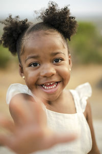 Optimistic little black girl in white clothes with hair buns looking at camera with smile on blurred background of nature