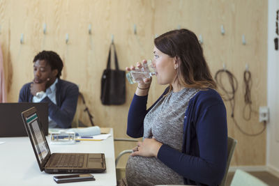 Pregnant businesswoman drinking water while sitting with laptop in meeting