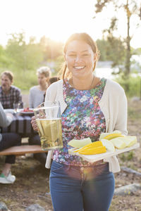 Portrait of happy woman holding beer jug and muskmelon slices with friends sitting at picnic table in background