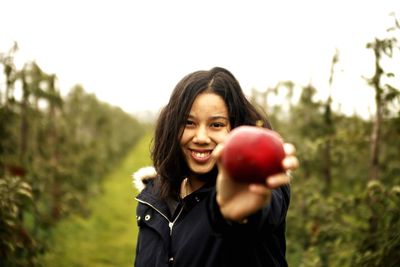 Portrait of smiling young woman holding apple against sky