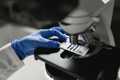 Crop anonymous scientist in protective gloves putting sample under microscope lens while conducting chemical research in laboratory