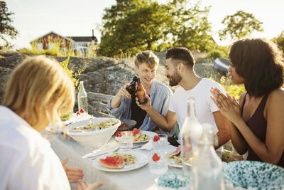 Men toasting beer bottles while sitting with friends at picnic table