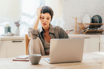 Young brunette woman sitting in the kitchen at the workplace uses a laptop for communication