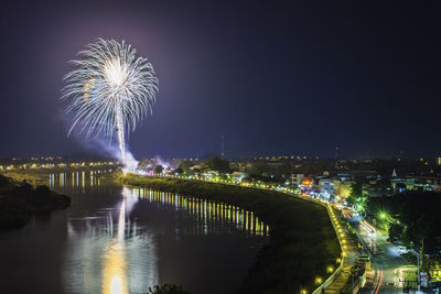 Firework display over river by illuminated city against sky at night