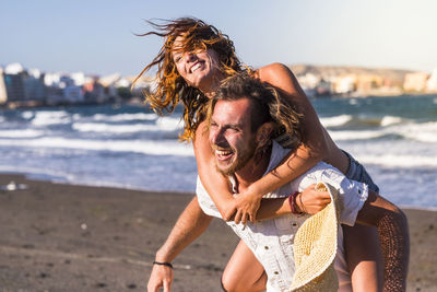 Man piggybacking cheerful woman at beach against sky