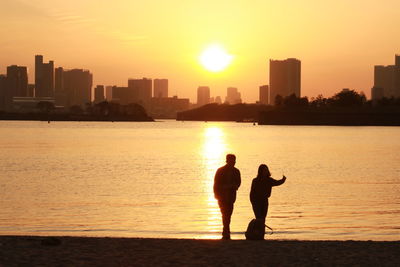 Silhouette friends standing on sea by cityscape against sky during sunset