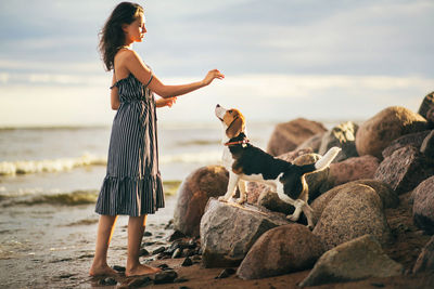 Side view full length of young woman standing by beagle at beach