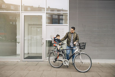 Smiling father riding bicycle with son sitting in safety seat on sidewalk in city