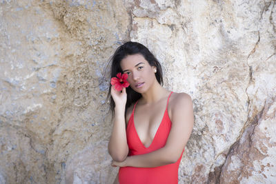 Side view of a young woman in a red swimsuit standing by a rock wall looking at the camera