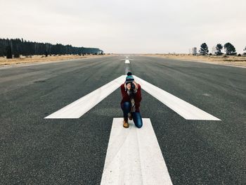 Young man photographing while kneeling on road against sky