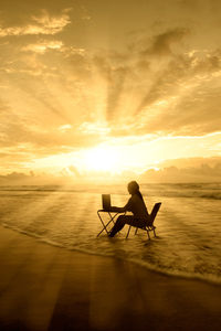 Silhouette woman using laptop while sitting on chair at beach during sunset