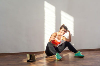 Full length of woman gesturing while sitting against wall at gym