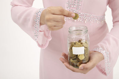 Midsection of woman holding coin over glass jar against white background